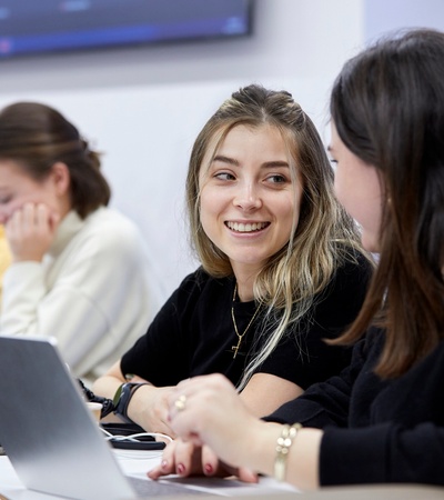 A group of students engaged in conversation and work at a table in a classroom setting.