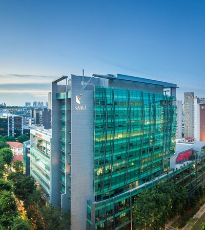 The image shows a modern glass building with greenery surrounding it in an urban setting during dusk.