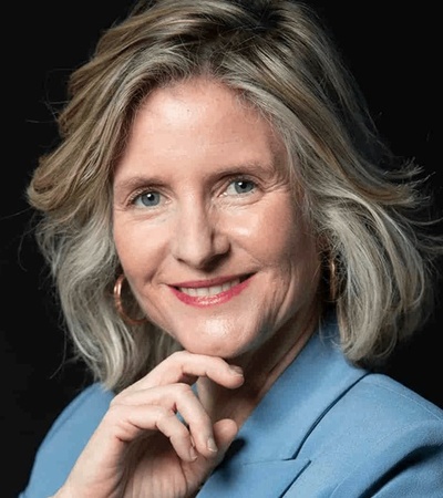 A portrait of a smiling woman with gray hair wearing a blue blazer against a black background.