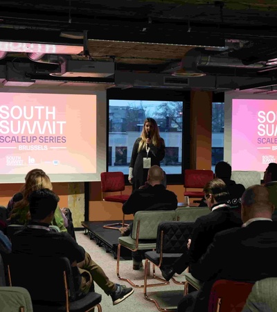 A woman is presenting at a conference with a large screen displaying the event's logo.