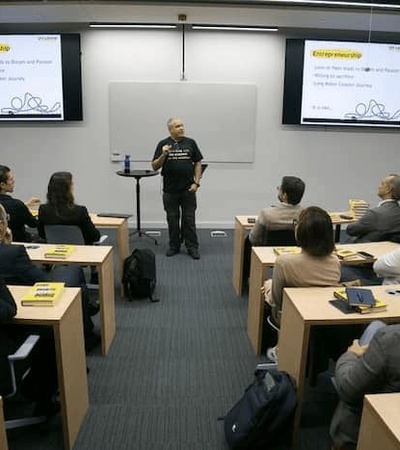 A person presenting in front of an attentive audience in a seminar room with slides displaying the topic Entrepreneurship.