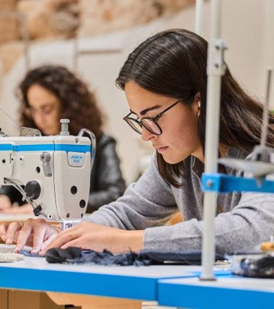 A woman in glasses is focused on sewing fabric with a sewing machine at a workshop, with another person visible in the background.