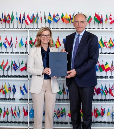 A formal meeting captured in a photograph with two individuals holding a document in front of a wall filled with flags.