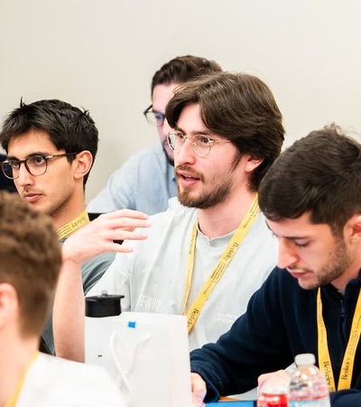 A group of young men engaged in a discussion at a conference, with one speaking and the others listening.