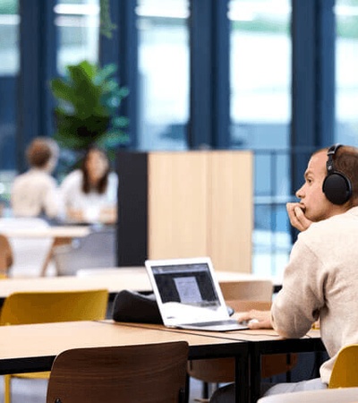 A man wearing headphones is using a laptop in a modern office space with other people in the background.