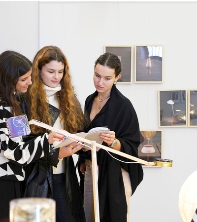 Three women examining documents in an art gallery with photos displayed on the wall.