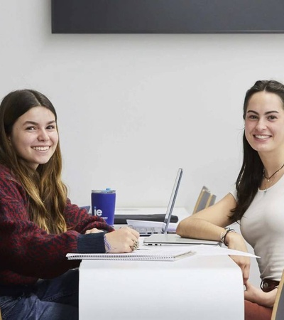 Three women smiling at a table in a classroom setting with laptops and notebooks.