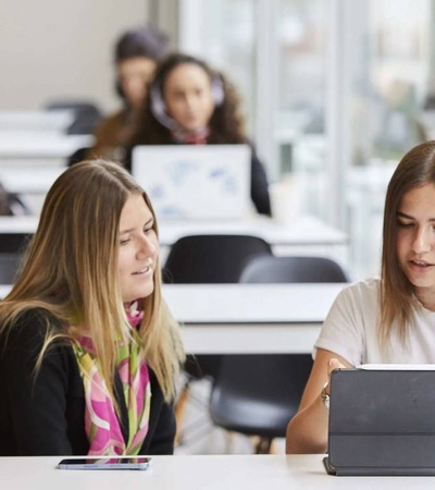 Two women collaborating on a work project with laptops at a white desk in an office environment.