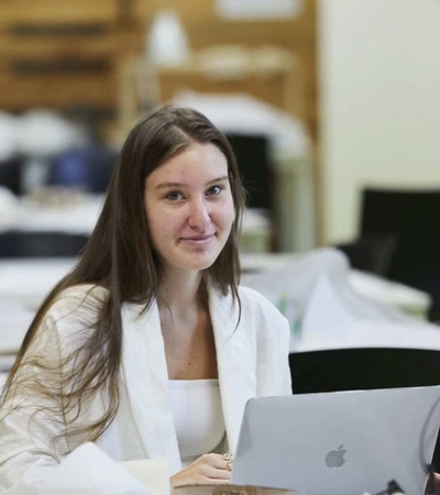 A young woman sits at a desk in an office environment, working on a laptop.