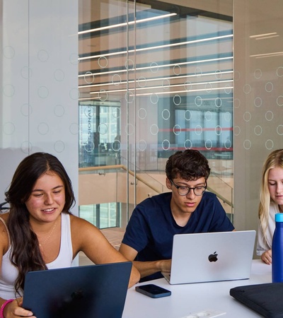 Three students are collaborating at a table with laptops in a modern study environment.