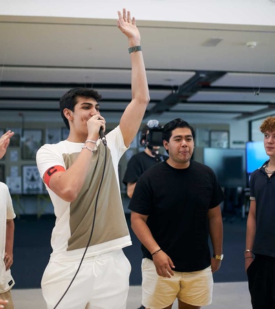 A young man enthusiastically singing into a microphone surrounded by cheering friends in a casual indoor setting.
