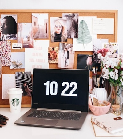 A stylish and creative workspace featuring a laptop displaying the time, surrounded by decorative items and images on a cork board.