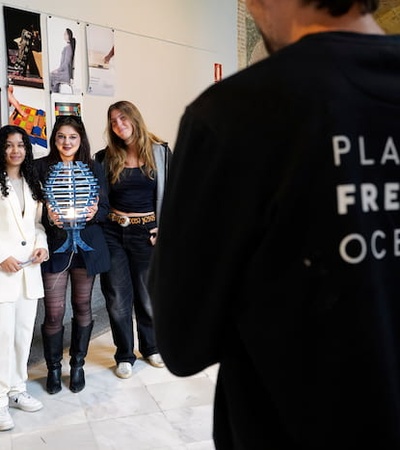 Three individuals pose for a photo at a display featuring various artistic works, while another person in a 'Plastic Free Oceans' shirt is seen from the back.