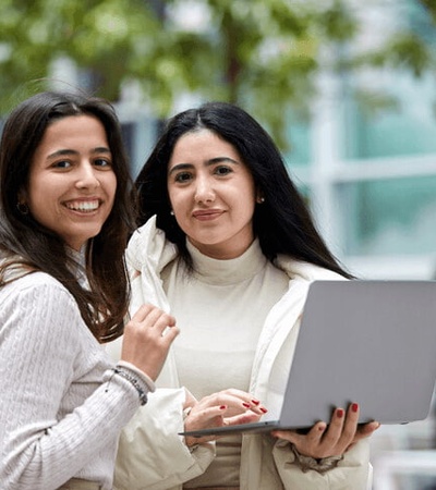 Two women holding a laptop and smiling outside a modern building.
