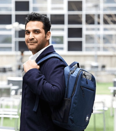 A man in a business suit with a backpack standing outside with tables and chairs in the background.