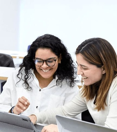 Two women are smiling and interacting with each other over a tablet in a modern office setting.