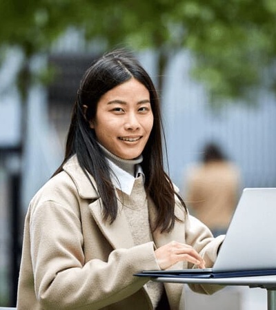 A young woman is smiling while using a laptop at an outdoor table.