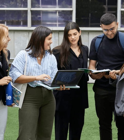 A group of five young adults are standing outdoors on grass, engaging in a discussion over a laptop and documents.