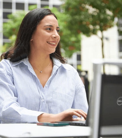 A woman smiles while using a laptop at an outdoor table in an urban setting.