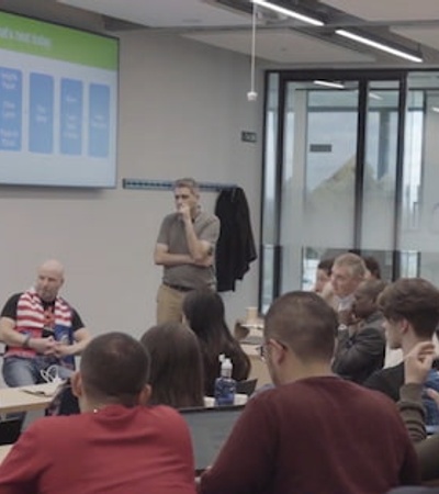 A group of people attending a lecture in a classroom with a senior presenter speaking in front of a digital presentation screen.