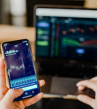 A person is checking stock market data on their smartphone while viewing a trading interface on a laptop.