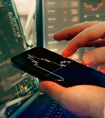 A hand is interacting with a smartphone displaying financial data, while a computer screen shows stock charts in the background.