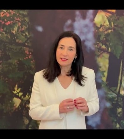 A woman in a white blazer stands in front of a forest backdrop, speaking and gesturing with her hands.