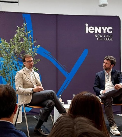 A discussion event at a New York college featuring two speakers seated on stage.