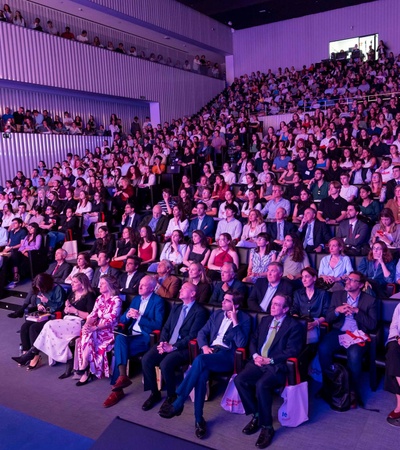 A large audience seated in a modern auditorium watching a presentation, illuminated by ambient blue and pink lighting.