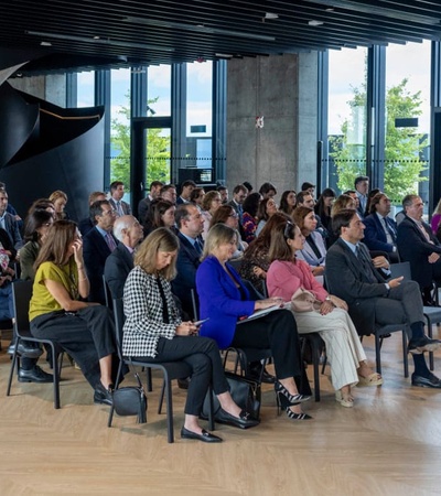 A group of people seated in rows at an indoor event, facing a focal point not visible in the image.