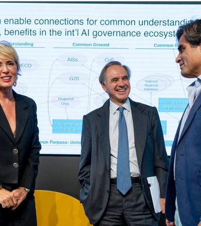 Three professionals are smiling and interacting in a seminar environment, with a backdrop displaying a presentation about legal frameworks and IE University's branding.