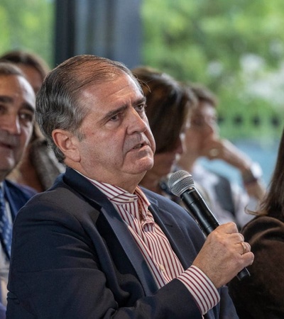 An older man holding a microphone speaks at a conference, surrounded by other attendees listening attentively.