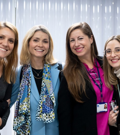 Four women smiling at the camera in an office-like environment with a white curtain in the background.