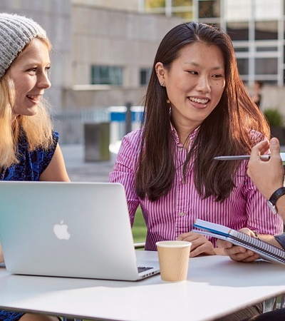 Three IE students speaking outside sitting on a table with a computer