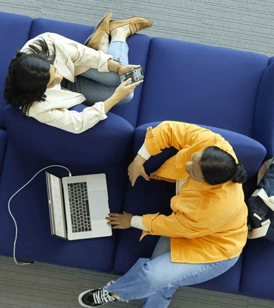 Two people sitting on a blue sofa, one working on a laptop and the other using a smartphone.