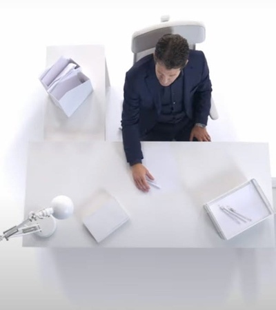 A person sitting at a minimalist white desk with papers and a lamp.