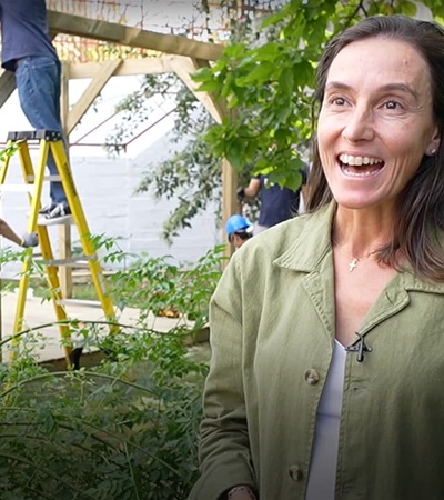 A smiling woman in a green jacket stands in a garden while workers in the background install a structure.