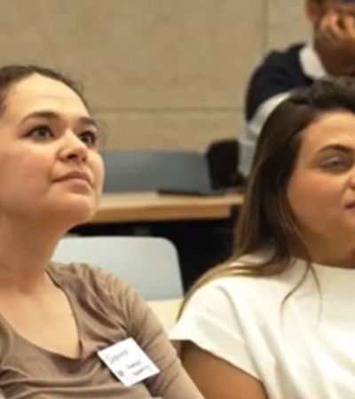 Two young women attentively listening in a lecture hall with other students around.