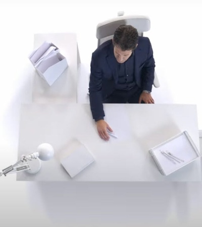 A man in a suit is seated at a minimalist white desk with documents and a lamp.