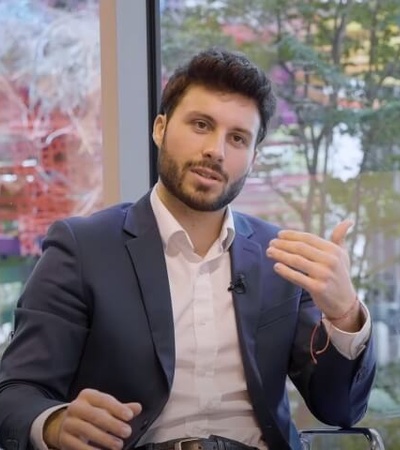 A man in a business suit is sitting and gesturing while talking, with a blurred office background visible through a window.