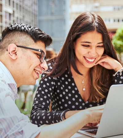 Two people, a man and a woman, are smiling and looking at a laptop together outdoors at a table in an urban setting.