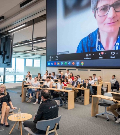A live virtual conference session taking place in a modern classroom with a large screen showing a remote participant.