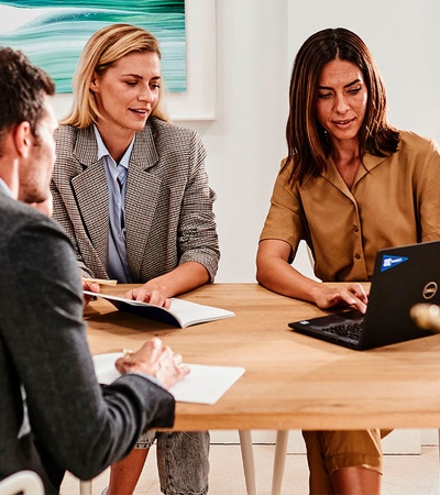 Three professionals, two women and one man, are engaged in a business meeting around a table, with one woman presenting on a laptop.