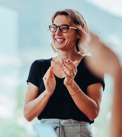 A woman with glasses clapping hands and smiling in a meeting or seminar room.