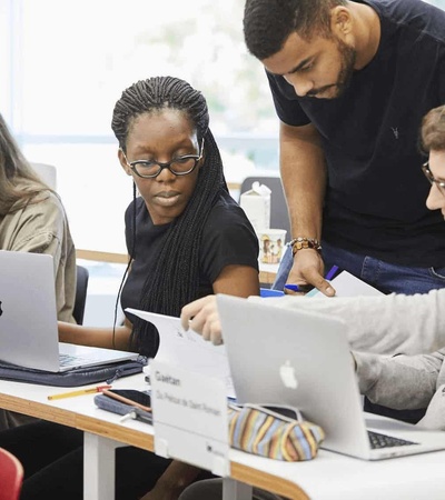 A group of students working together on laptops at a table in a classroom.