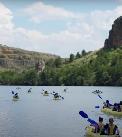 Groups of people are kayaking on a calm lake surrounded by rocky cliffs under a blue sky.