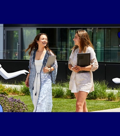 Two young women are conversing and walking with books in hand on a sunny campus, accompanied by illustrations of two cartoon-style figures.