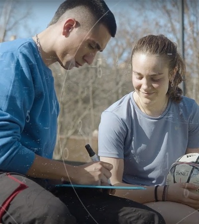 Two young people, one male and one female, are sitting outdoors on the grass with a basketball and a notebook, engaging in a conversation.