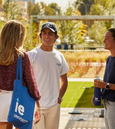 Three young adults are chatting happily on a university campus, holding reusable cups and a bag branded with the university logo.