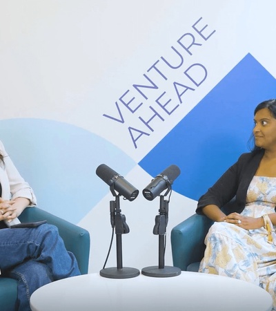 Two women are seated in front of microphones, discussing in a modern setting.